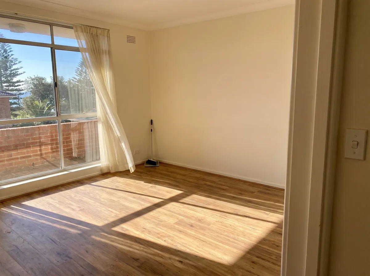 A small emptied Balgowlah walk-up flat living room, timber-look floor, a wide window with a sheer curtain and bright morning light, cleared and swept for the next tenant