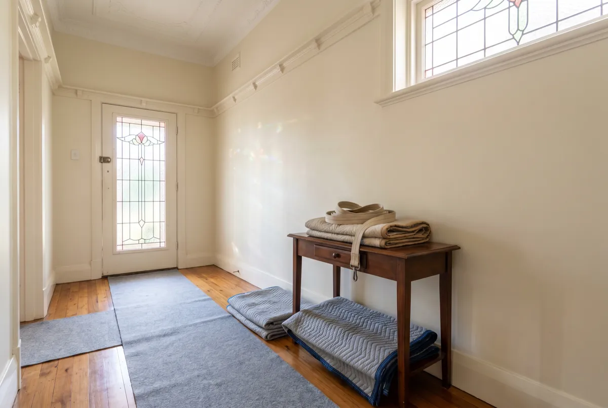 The entry hall of a 1930s Federation home during a careful clearance: original polished floorboards laid with clean felt runners and moving blankets, a period leadlight front door, a coiled carry strap resting on a hall table