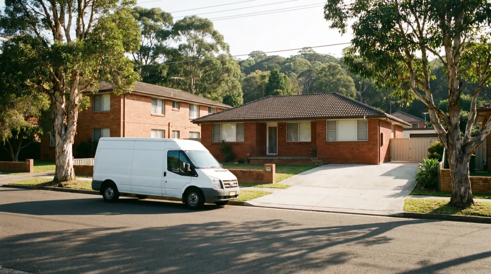 A quiet leafy Manly Vale street with a plain white van pulled up beside a brick home's wide driveway, a low-rise brick unit block next door and a green bushland ridge rising behind the rooftops