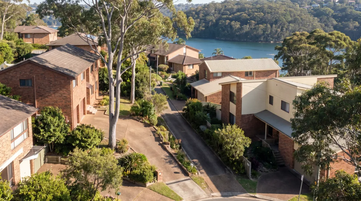 A steep Seaforth street of large brick and render family homes stepping down a terraced hillside toward the calm blue water of Middle Harbour, tall gum trees and long driveways