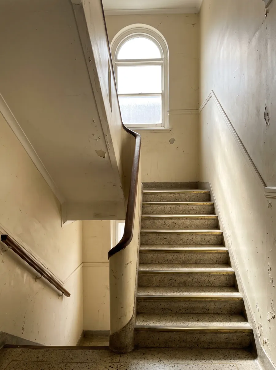 A cream art-deco stairwell with terrazzo stairs turning at a half-landing, lit from a tall window